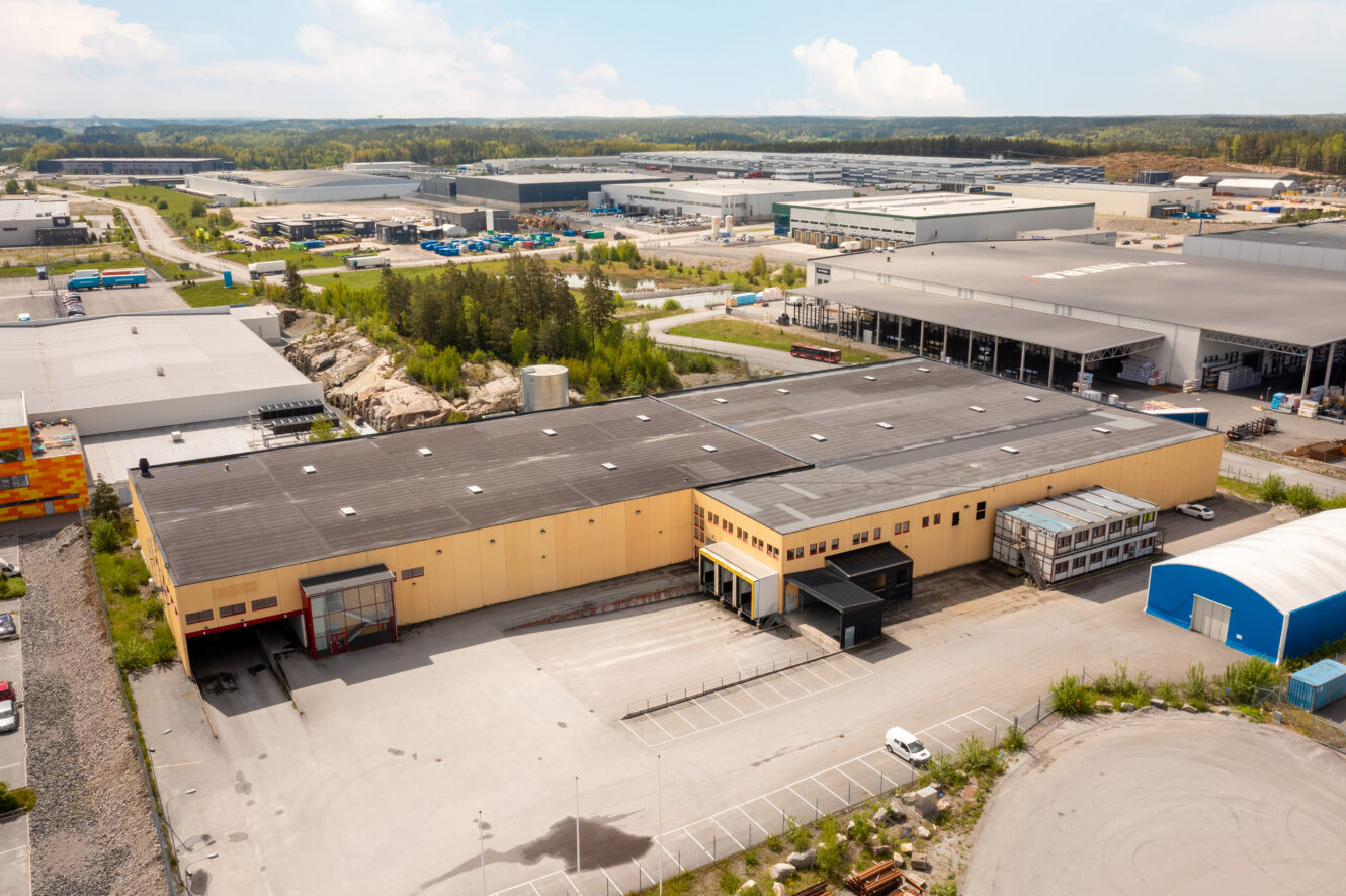 Flygfoto över en stor industrihall med gul fasad i ett kommersiellt område, omgiven av parkeringsplatser, träd och andra industribyggnader.