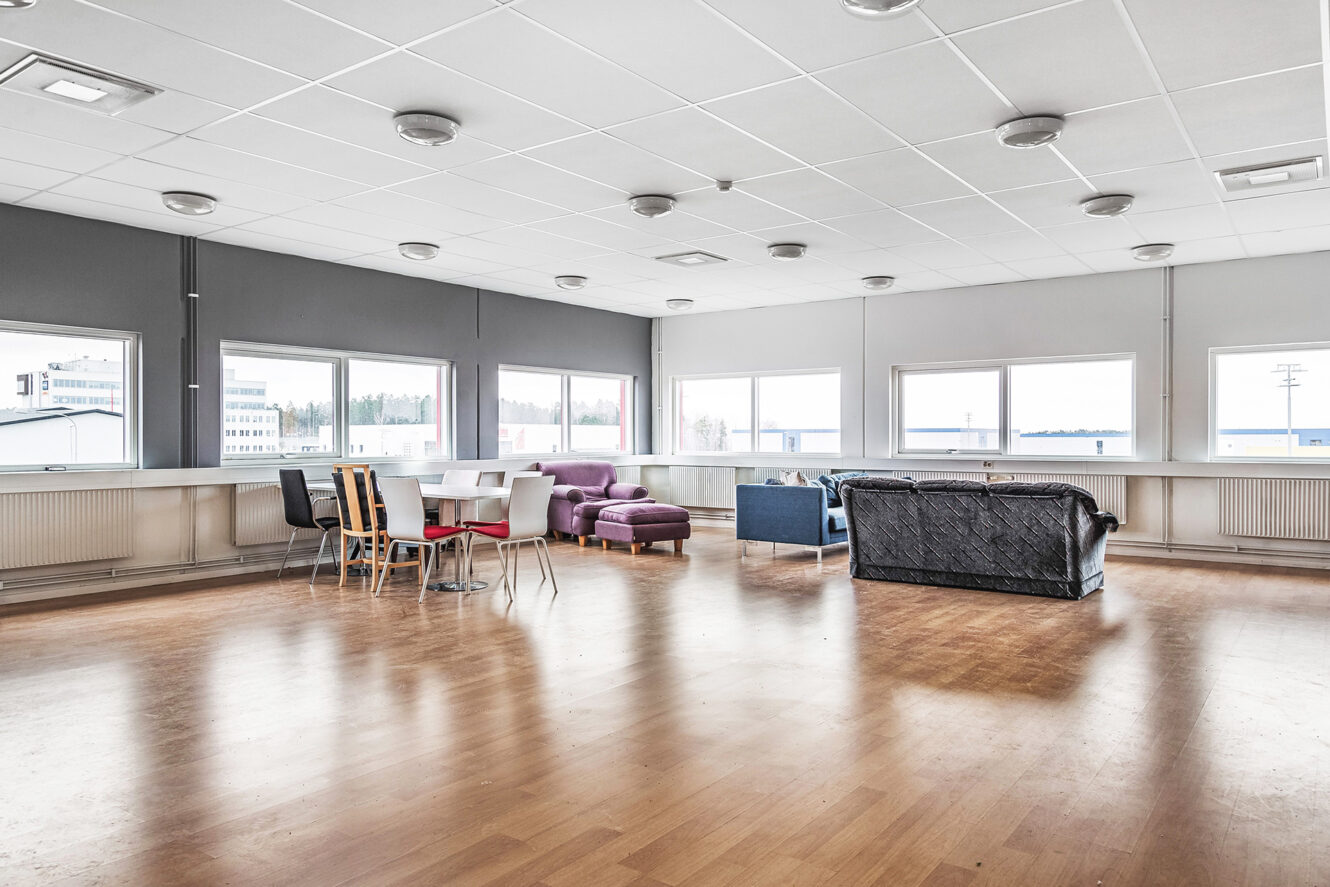 Ett rymligt, ljust rum med stora fönster, trägolv, olika stolar, ett matbord och flera omaka soffor placerade nära väggarna.