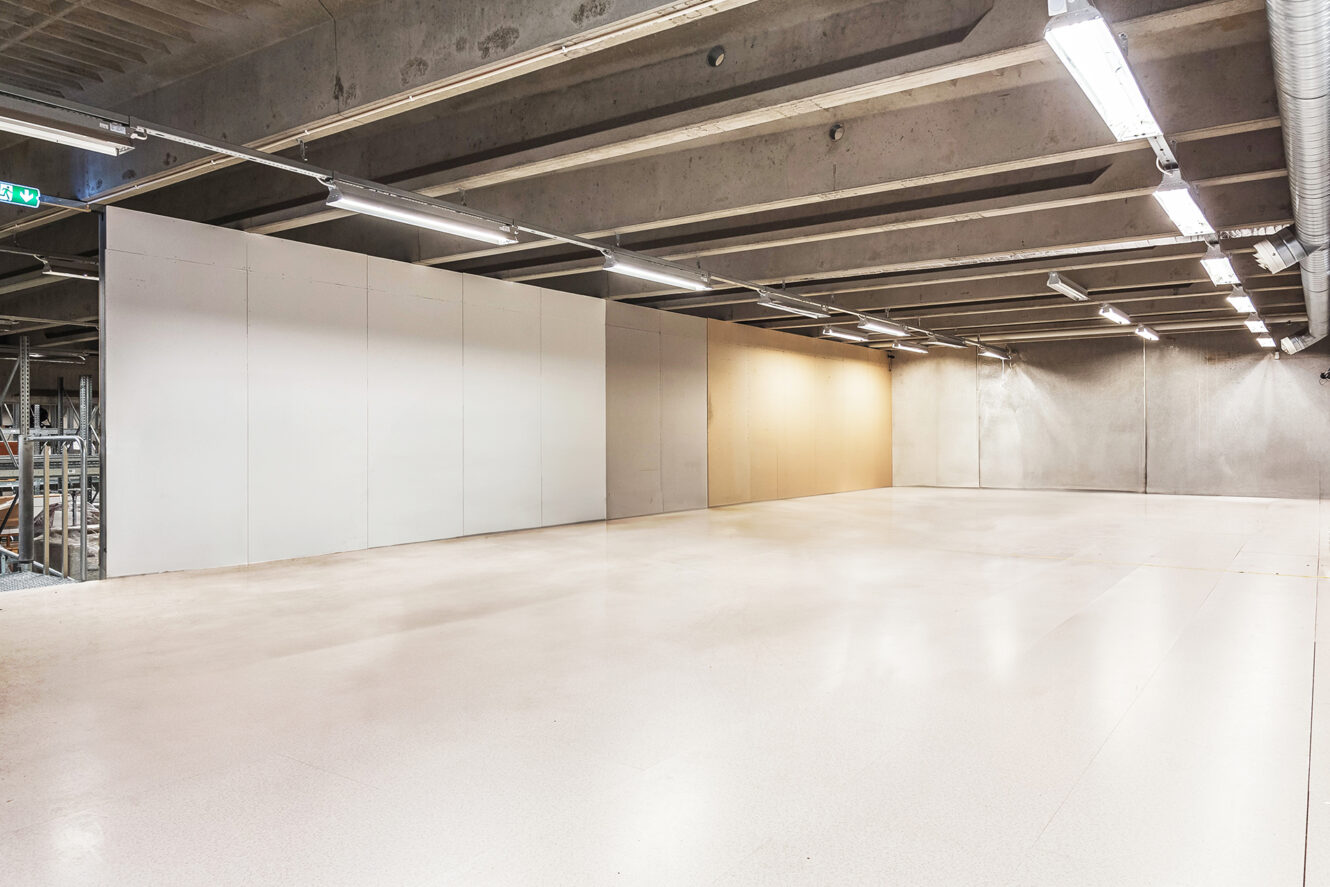 Ett stort, tomt industrirum med betongväggar, synliga takbjälkar och stark takbelysning. Hyllor syns längst till vänster.