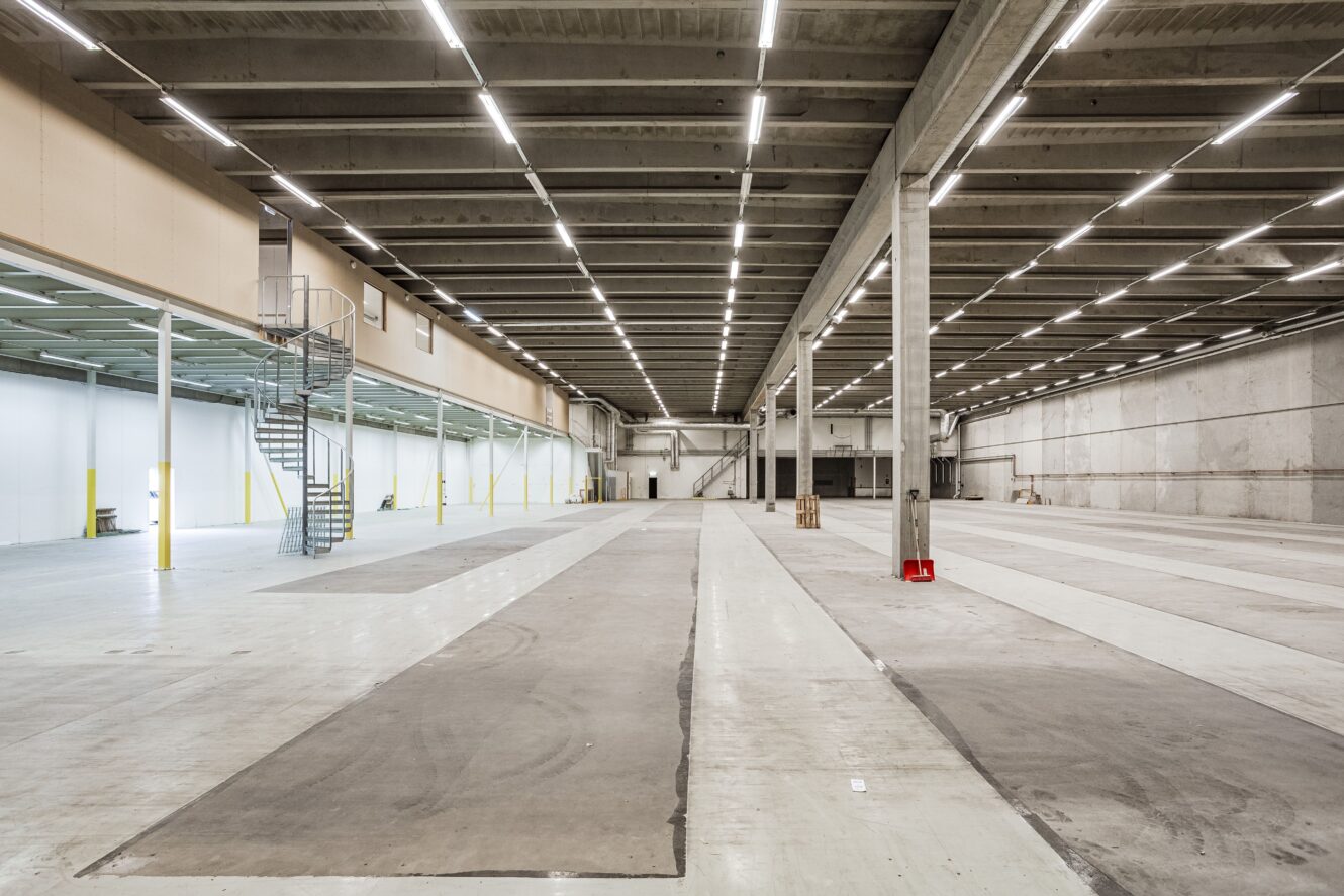 Stor, tom lagerlokal med högt i tak, synliga bjälkar, betonggolv och en spiraltrappa av metall till vänster. Utrymmet är upplyst av en stark takbelysning.