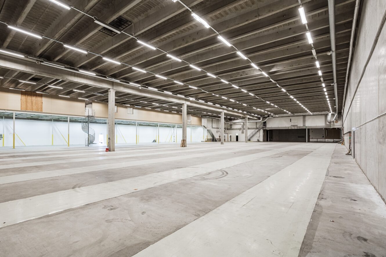 Stor, tom lagerlokal med betonggolv, högt i tak, synliga bjälkar och stark takbelysning. Det finns en spiraltrappa i metall längs ena väggen.