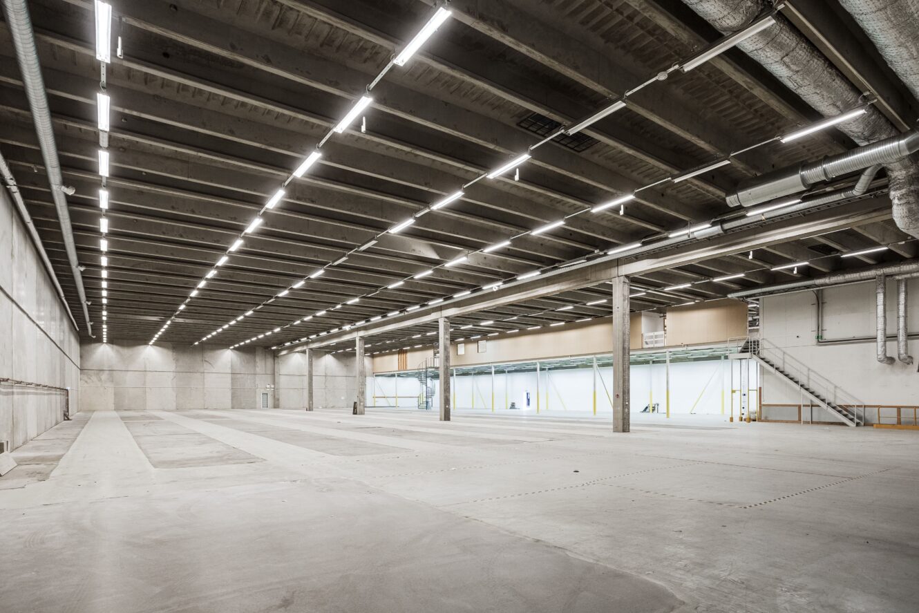 Stort, tomt industrilager med betonggolv, högt i tak, synliga bjälkar och stark takbelysning. Väggarna är släta och det finns lastkajer som syns på ena sidan.