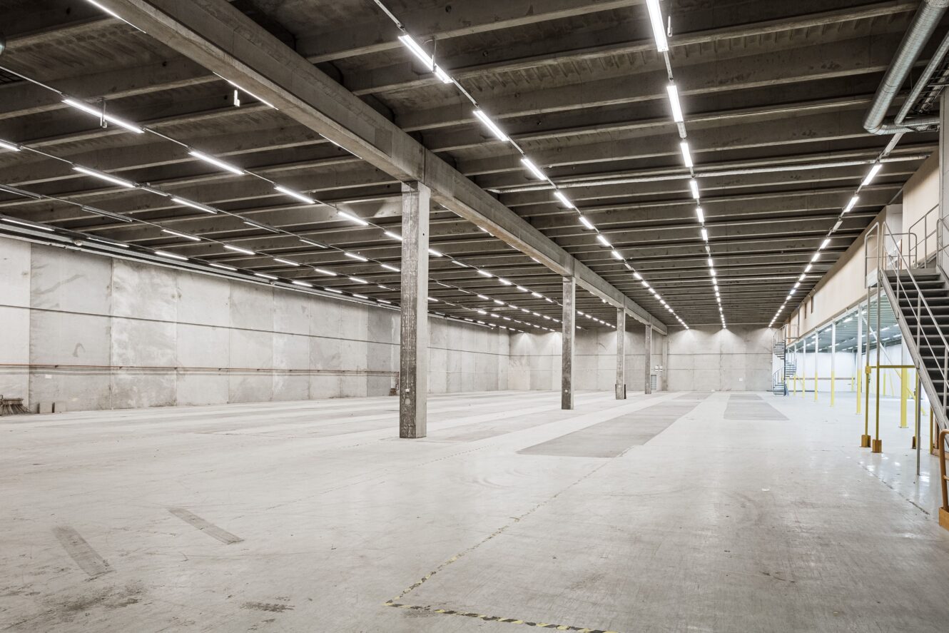 Stor, tom lagerlokal med betonggolv, högt i tak, synliga bjälkar och rader av taklampor; metalltrappa på höger sida.