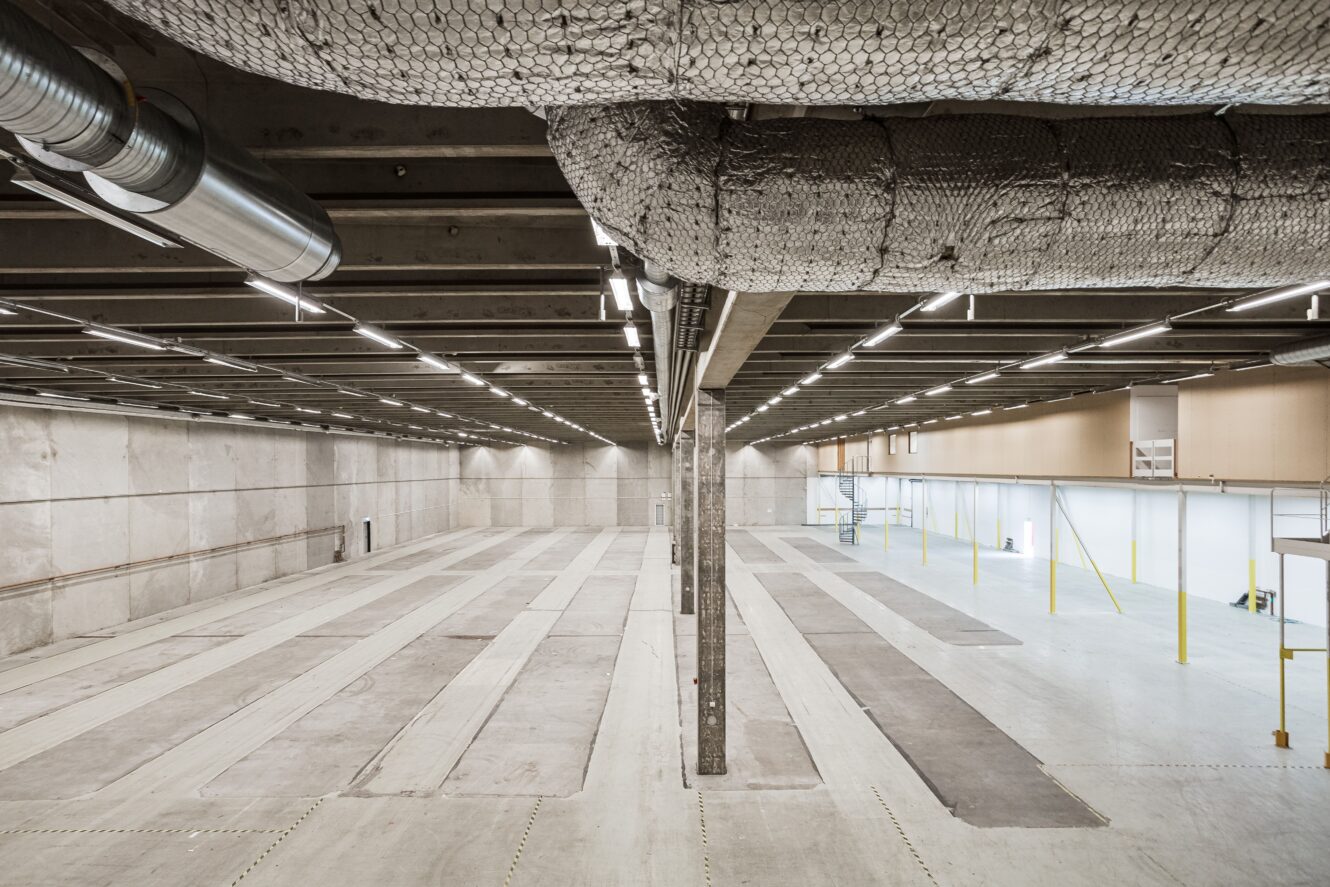 Stort, tomt industrilager med synliga kanaler, högt i tak, betonggolv och stark takbelysning. Gula stolpar och mezzanin syns nära den bortre väggen.