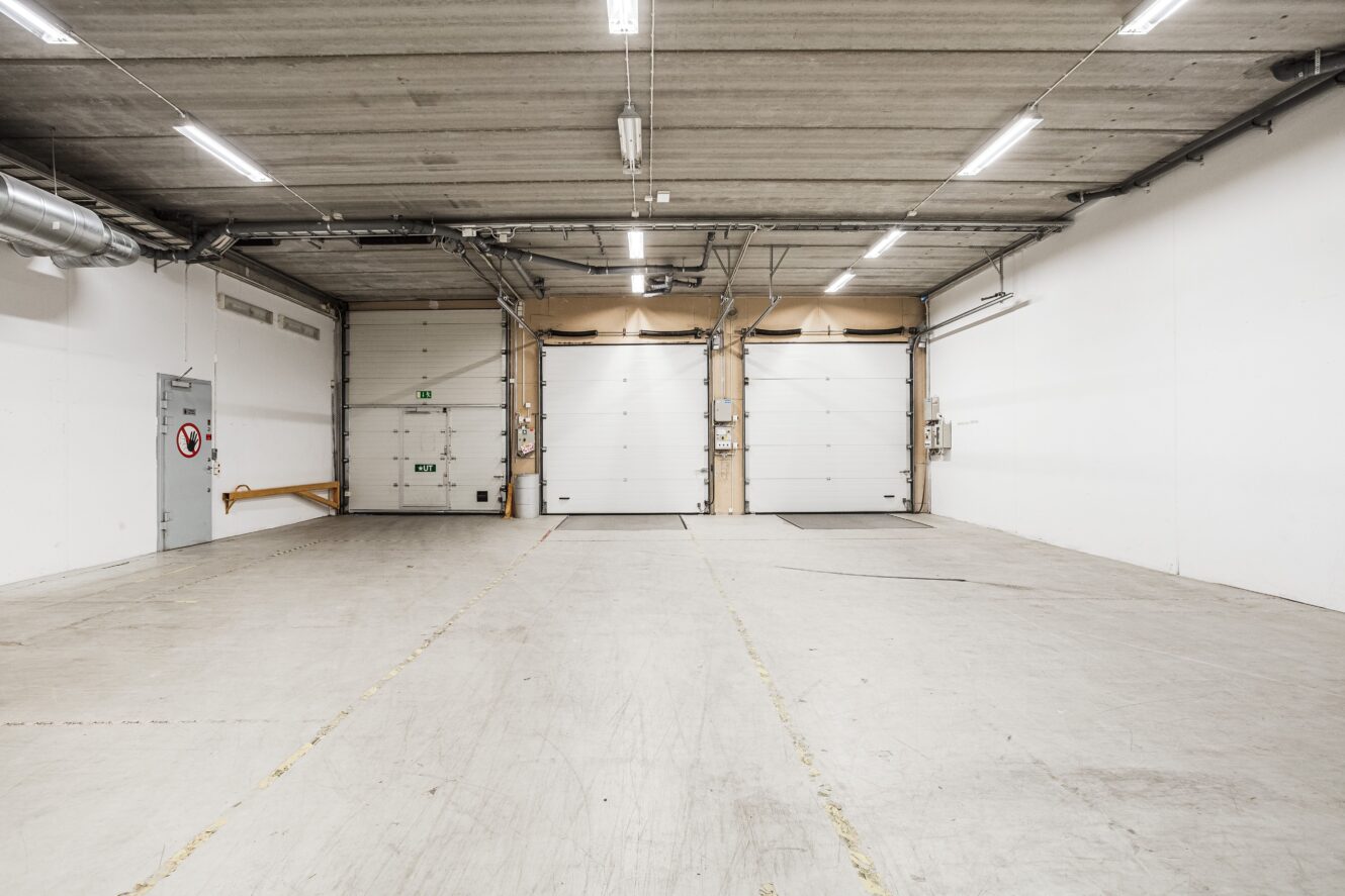 Tom lagerlokal med tre stora vita dörrar till lastkajer, takbelysning, vita väggar och betonggolv.