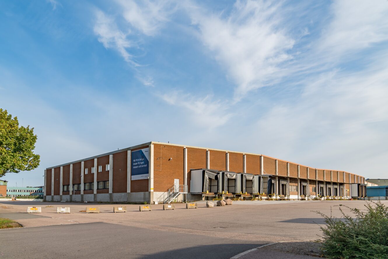 En stor, rektangulär lagerlokal i tegel med flera lastkajer och en i stort sett tom parkeringsplats under en delvis molnig himmel.