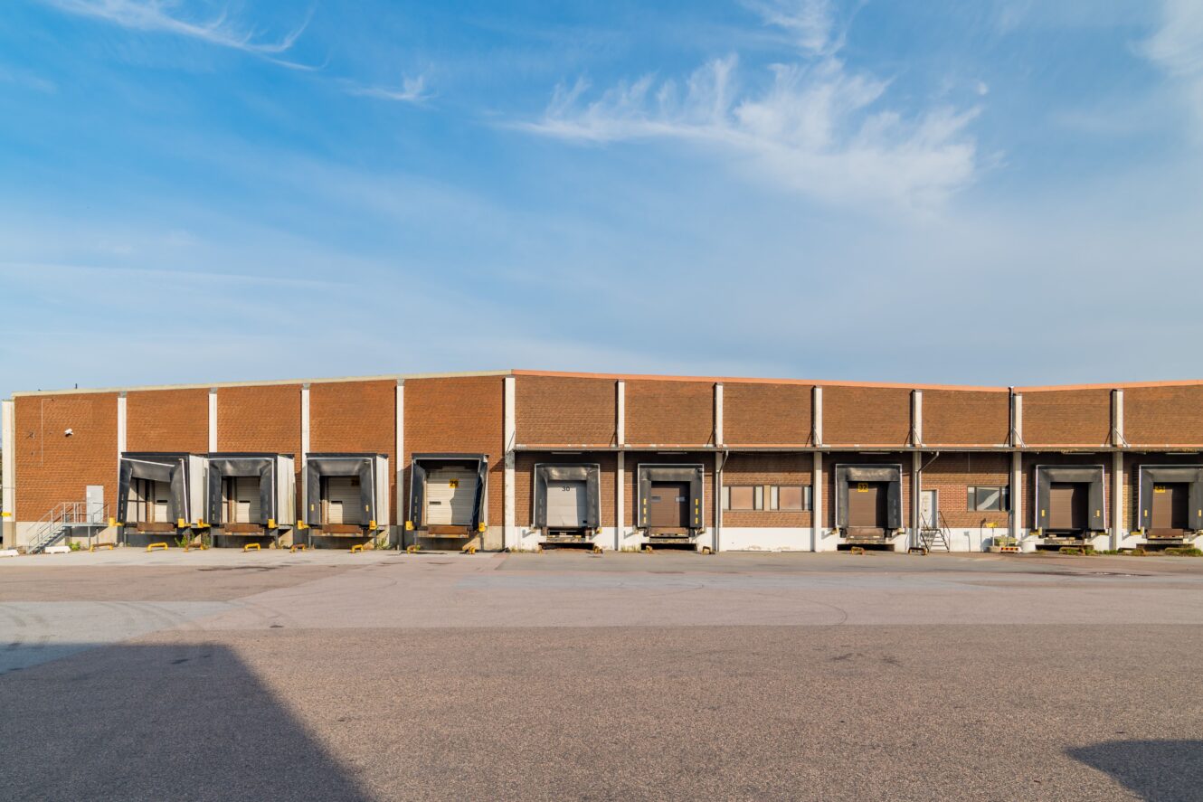 En rad lastkajer i anslutning till en stor brun industrihall under en delvis molnig himmel.