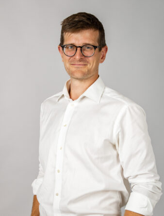 En man med glasögon och vit skjorta står mot en ljusgrå bakgrund och tittar in i kameran med ett neutralt uttryck.