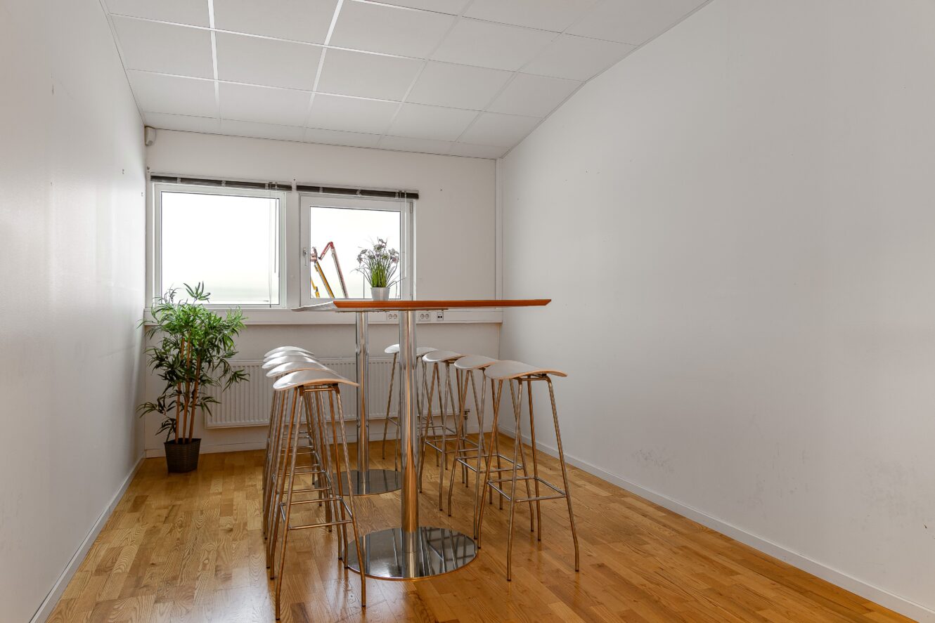 Ett litet rum med ett högt träbord, sex barstolar i metall, trägolv, vita väggar, två fönster, en krukväxt och en liten växt på fönsterbrädan.