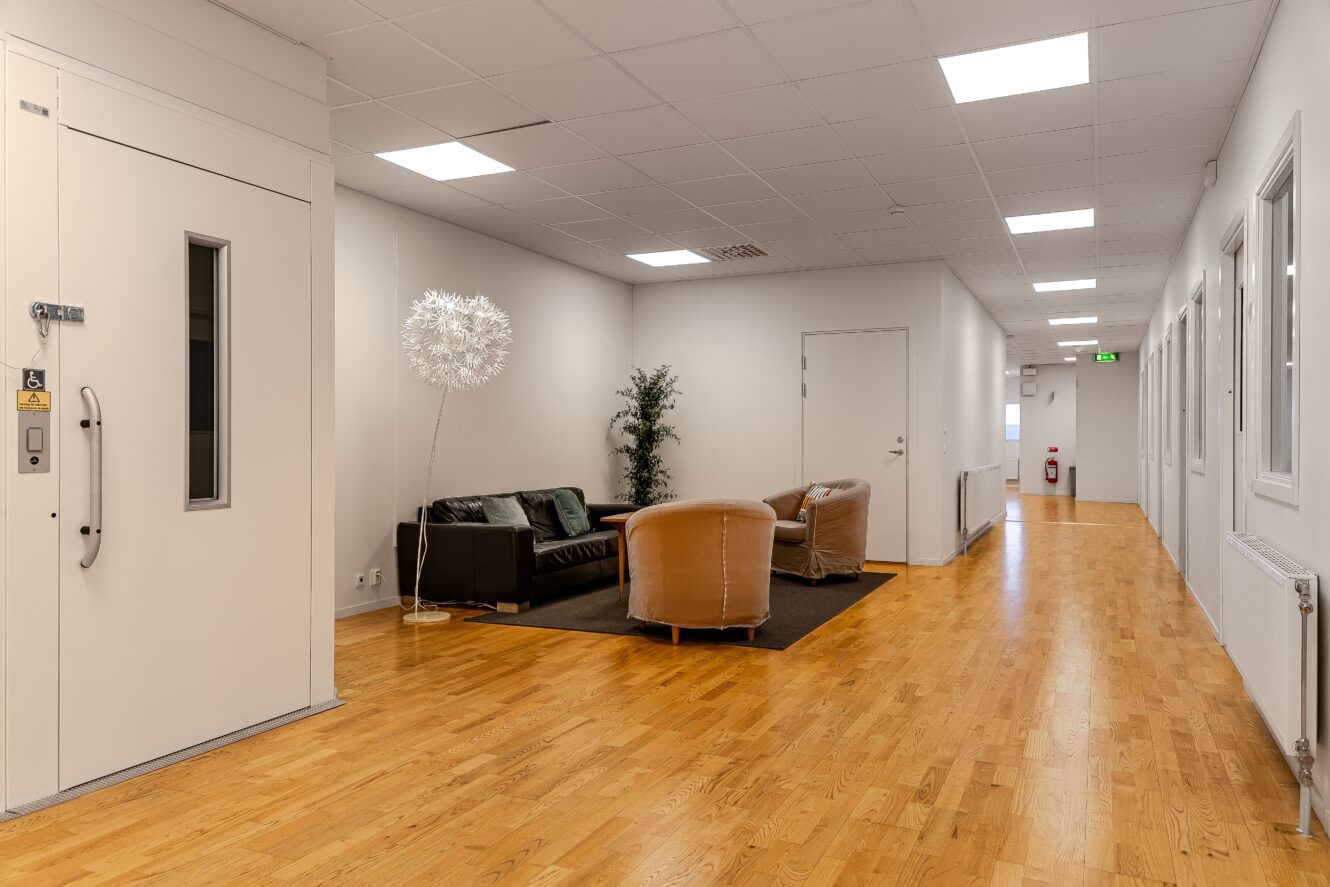 En hall med ljust trägolv, vita väggar, en hiss till vänster och en sittgrupp med soffor, fåtöljer och växter på en mörk matta.