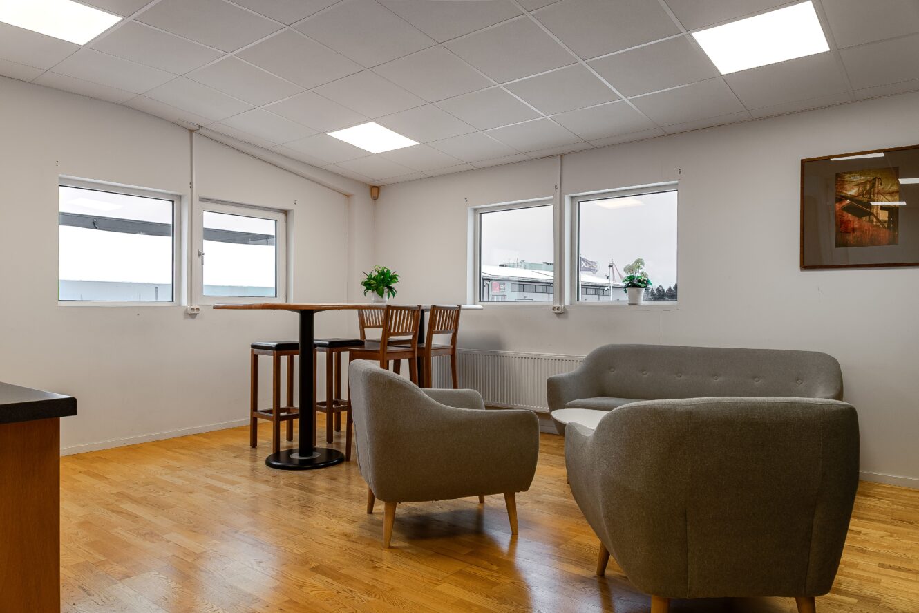 En modern lounge med två gråa fåtöljer, en matchande soffa, ett högt bord med stolar, trägolv och tre stora fönster.