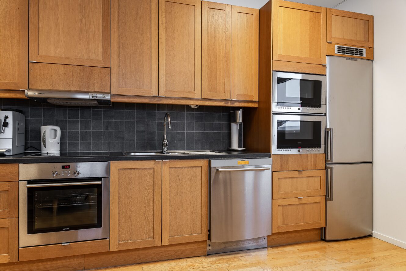 Modernt kök med träskåp, inbyggd ugn, vattenkokare, diskmaskin, dubbla väggugnar, kylskåp och svart kaklat stänkskydd.
