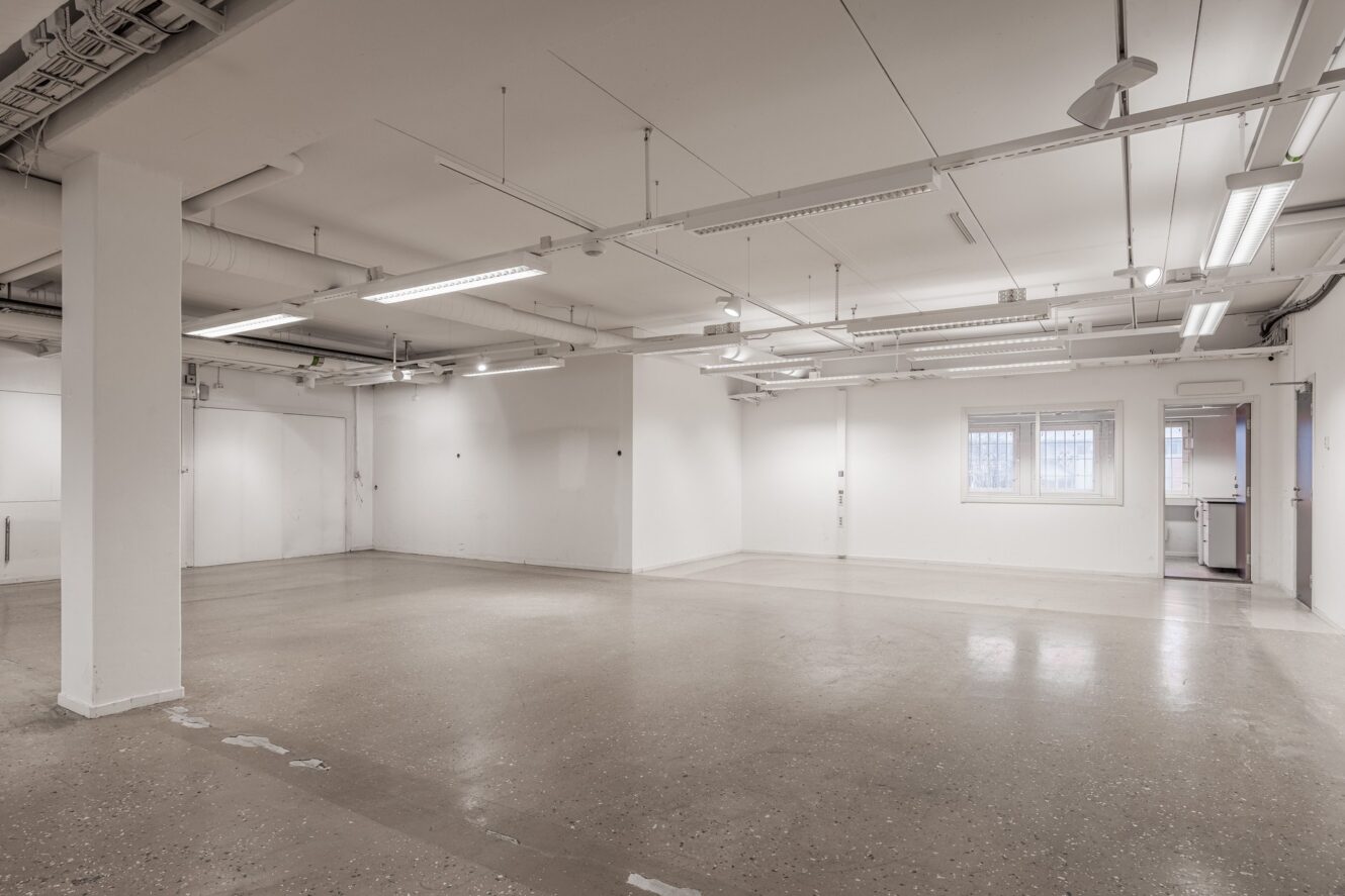 Tomt rum i industriell stil med vita väggar, synliga rör, taklampor, betonggolv, litet fönster och dörröppning till pentry.