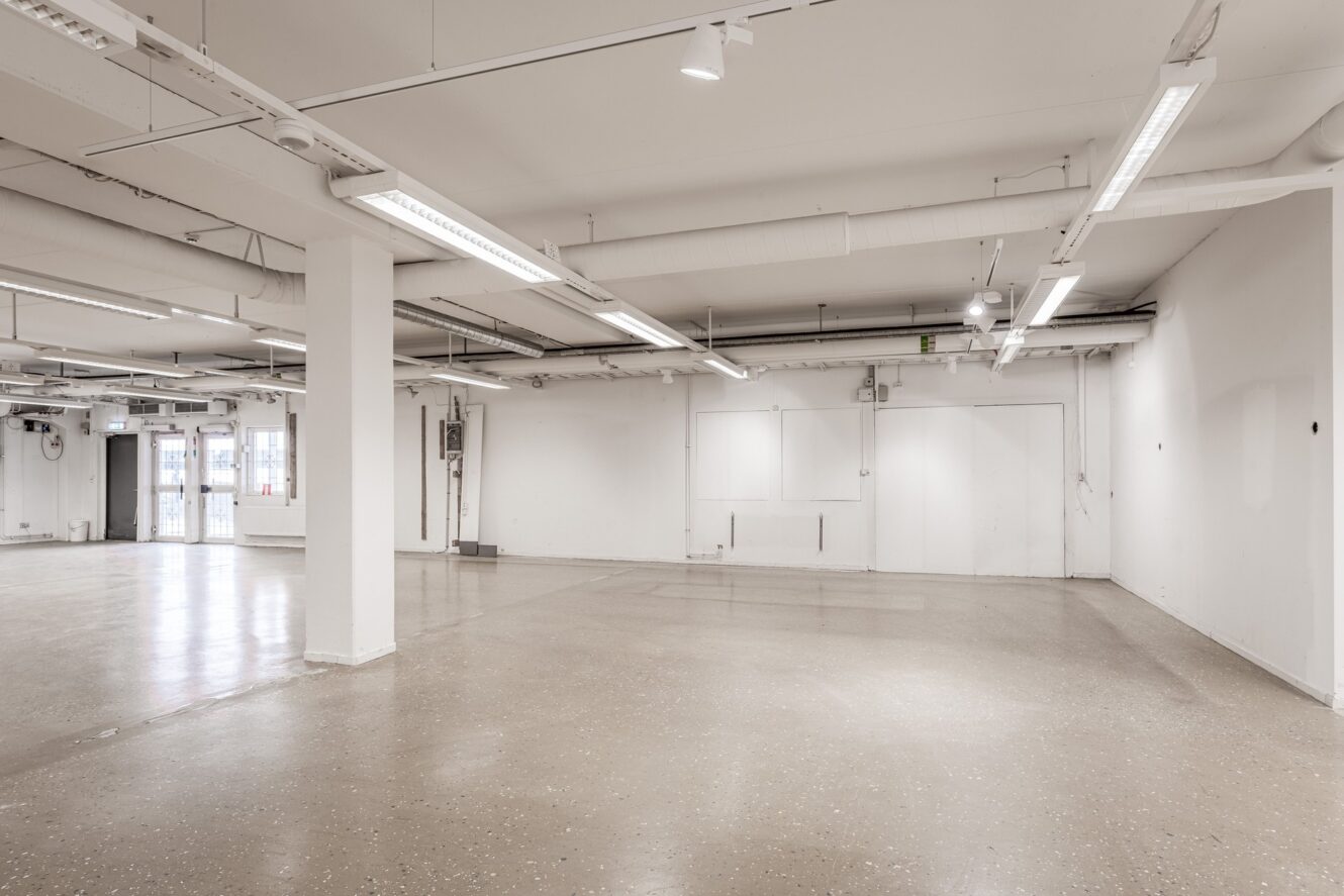 Rymlig, tom industrilokal med vita väggar, rör i taket, lysrör och ett polerat betonggolv.