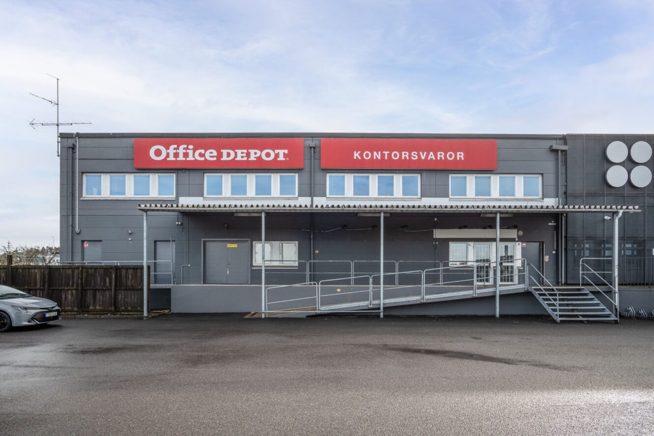 En grå affärsbyggnad med en Office Depot Kontorsvaror-skylt, flera fönster, en ramp, trappor och en tom parkeringsplats framför.