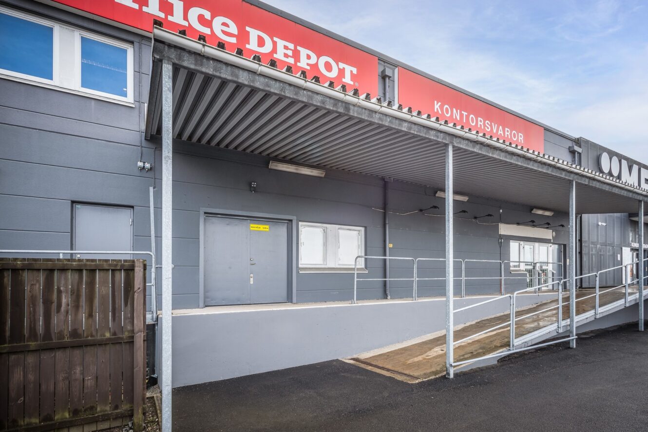 En grå affärsbyggnad med en lastkaj, metallramp och röd Office Depot-skylt. Scenen är tom under en delvis molnig himmel.