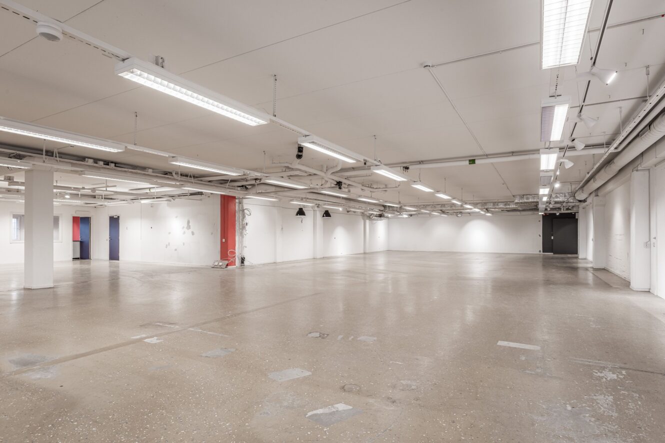 Stort, tomt industrirum med vita väggar, synliga rör i taket, lysrörsbelysning och polerat betonggolv.