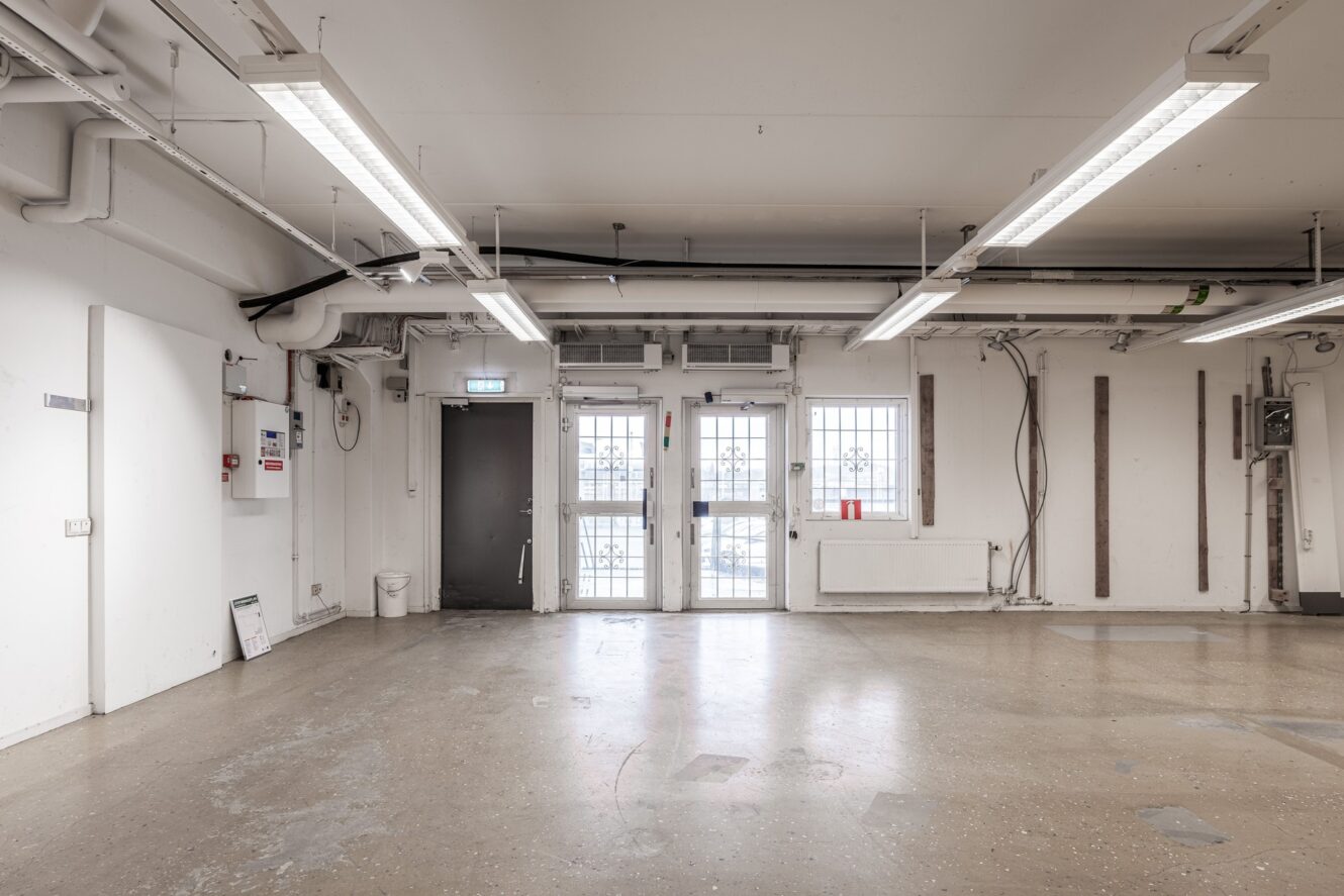 Tomt industriliknande rum med lysrör i taket, betonggolv, vita väggar, synliga rör, gallerförsedda fönster och en dörr på baksidan.