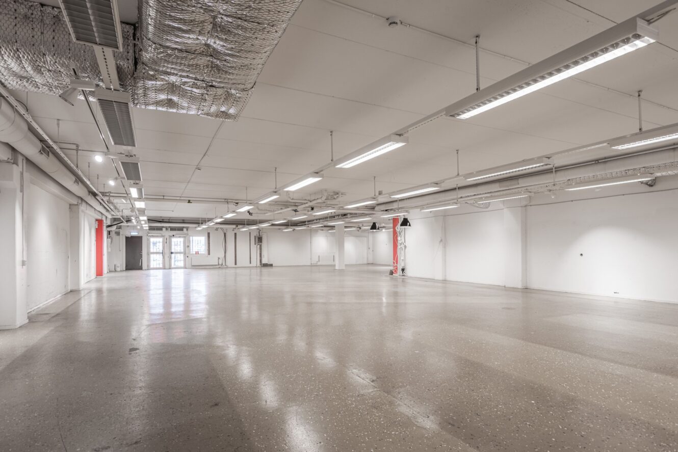 Stor tom industrilokal med vita väggar, synliga rör i taket, lysrör, betonggolv och röda dubbeldörrar i slutet.