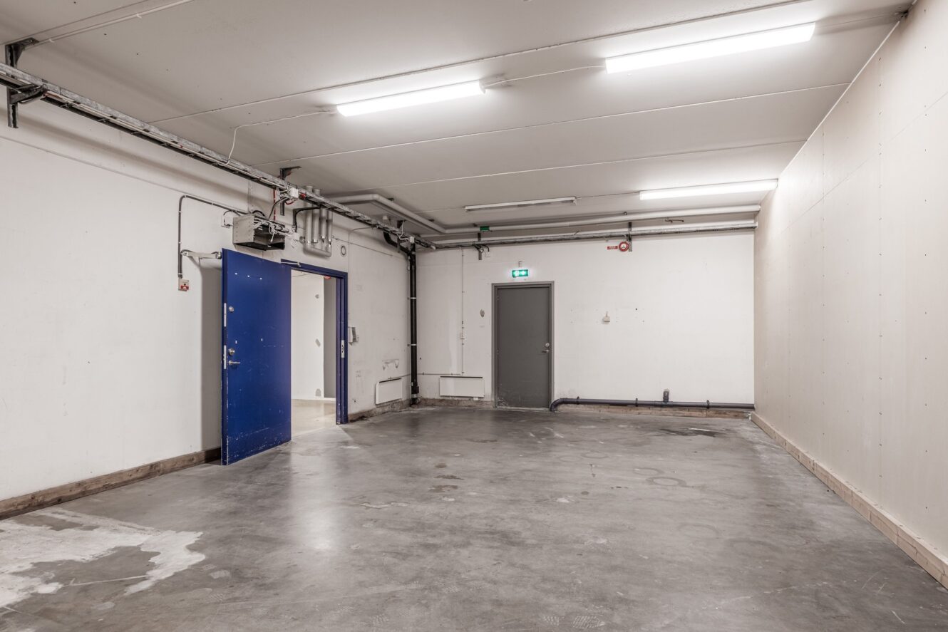 Ett tomt industrirum med betonggolv, vita väggar, starka taklampor, synliga rör och blå och grå metalldörrar.