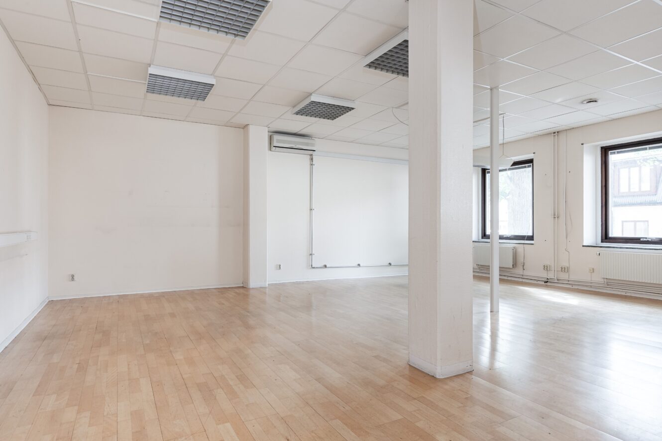 Tomt rum med ljust trägolv, vita väggar, en central pelare, stora fönster, taklampor och synlig HVAC-enhet.