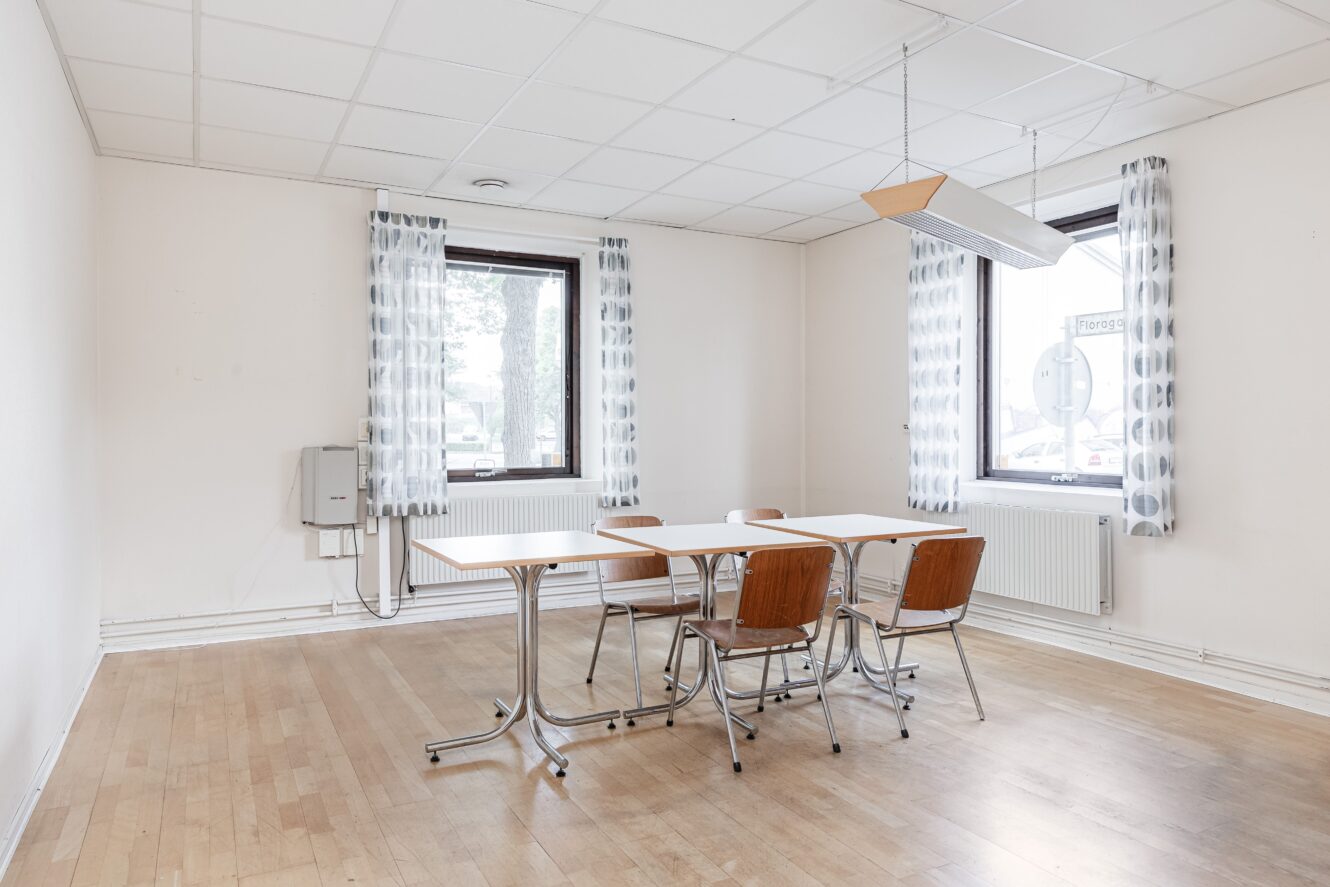 Ett litet, sparsamt möblerat rum med två fönster, vitt tak, trägolv, ett rektangulärt bord och fyra trästolar.