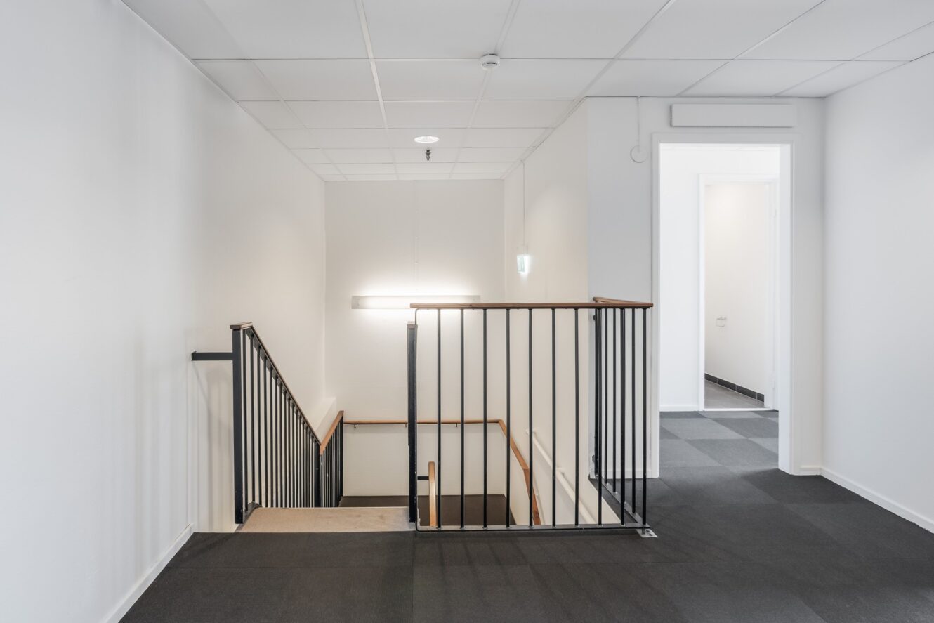 Ett modernt, minimalistiskt trapphus med svart räcke, vita väggar och en taklampa som leder till en lägre våning, bredvid en hall med heltäckningsmatta och en öppen dörröppning.