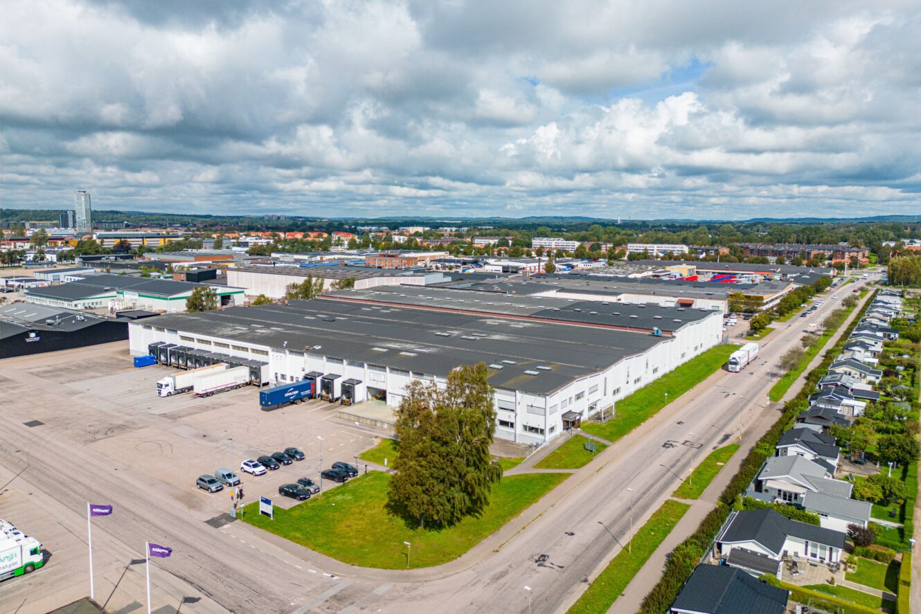 Ett stort industriellt lagerkomplex med lastkajer och lastbilar, omgivet av vägar, parkerade bilar och närliggande bostadshus under en delvis molnig himmel.