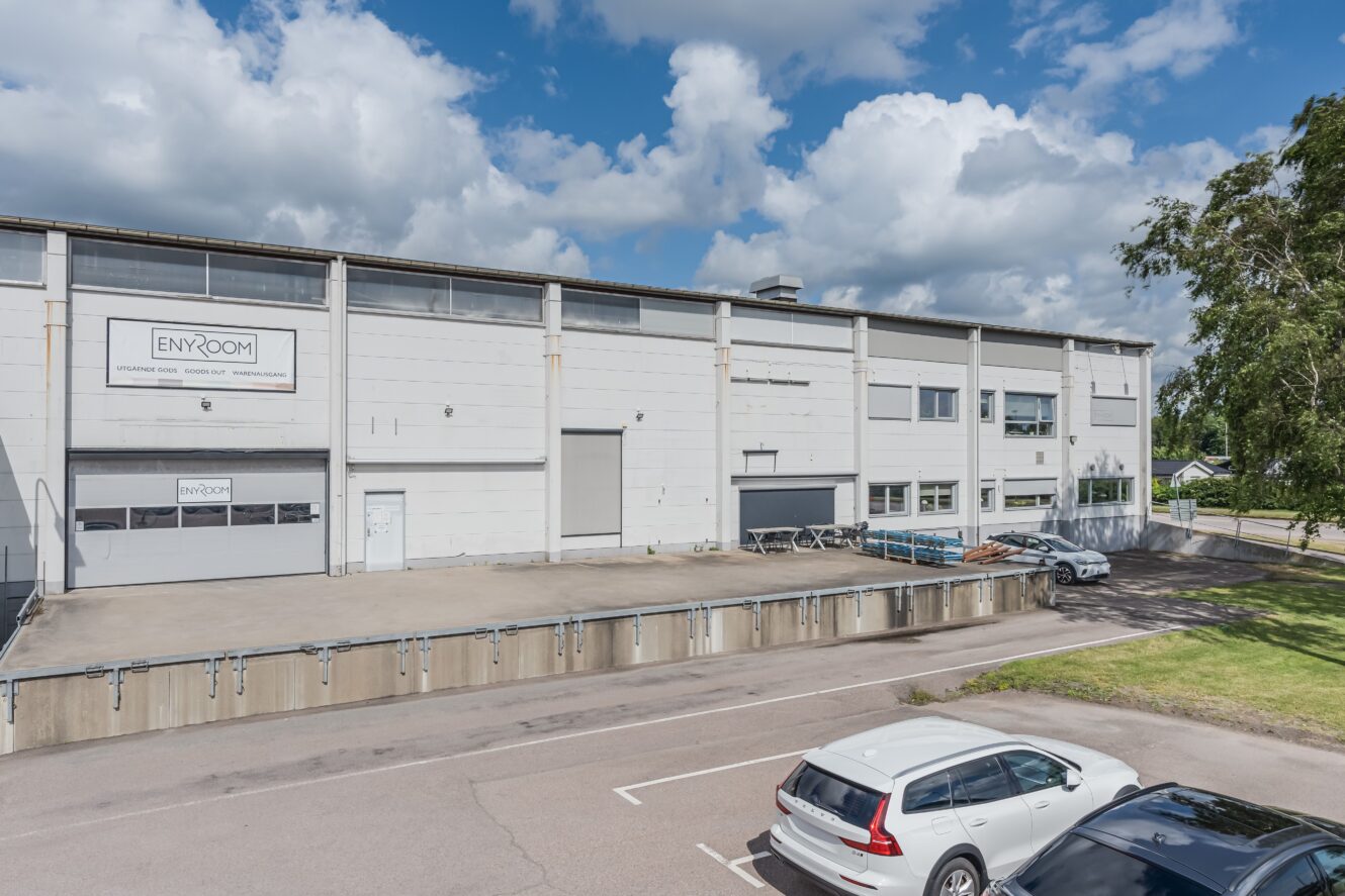 En vit industribyggnad med lastkajer och en företagsskylt, några parkerade bilar och en delvis molnig himmel ovanför.