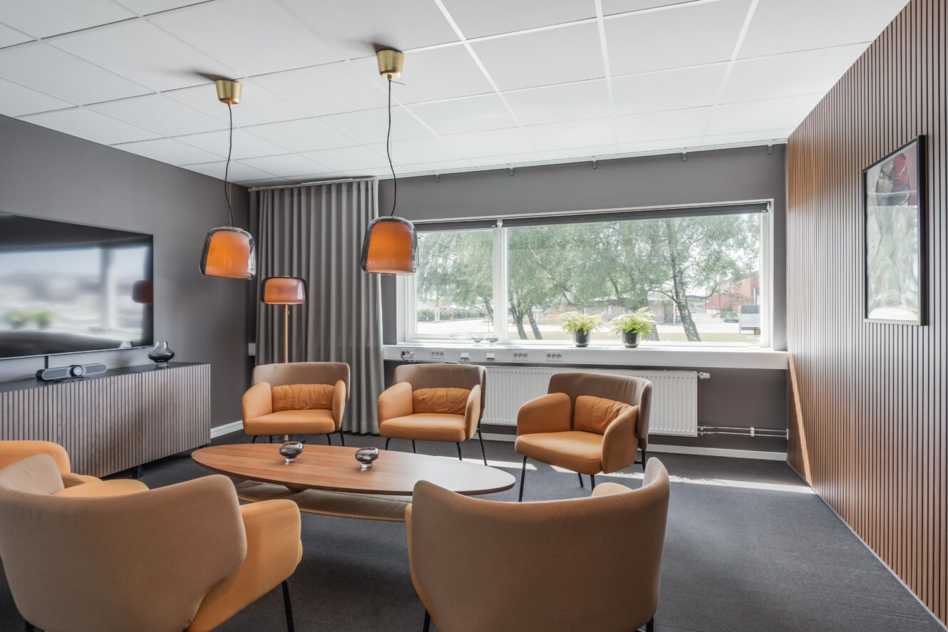 Modernt kontorsrum med solbruna fåtöljer, soffbord i trä, pendellampor, väggmonterad TV och ett stort fönster med krukväxter.