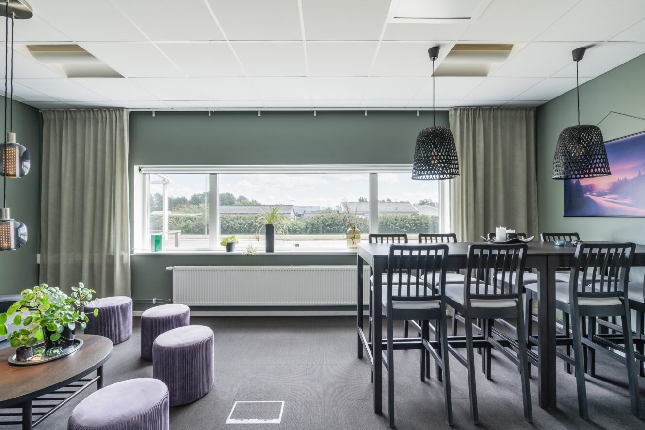 Modern kontorslounge med gröna väggar, ett långt fönster, högt bord med stolar, runda pallar, krukväxter och pendellampor.