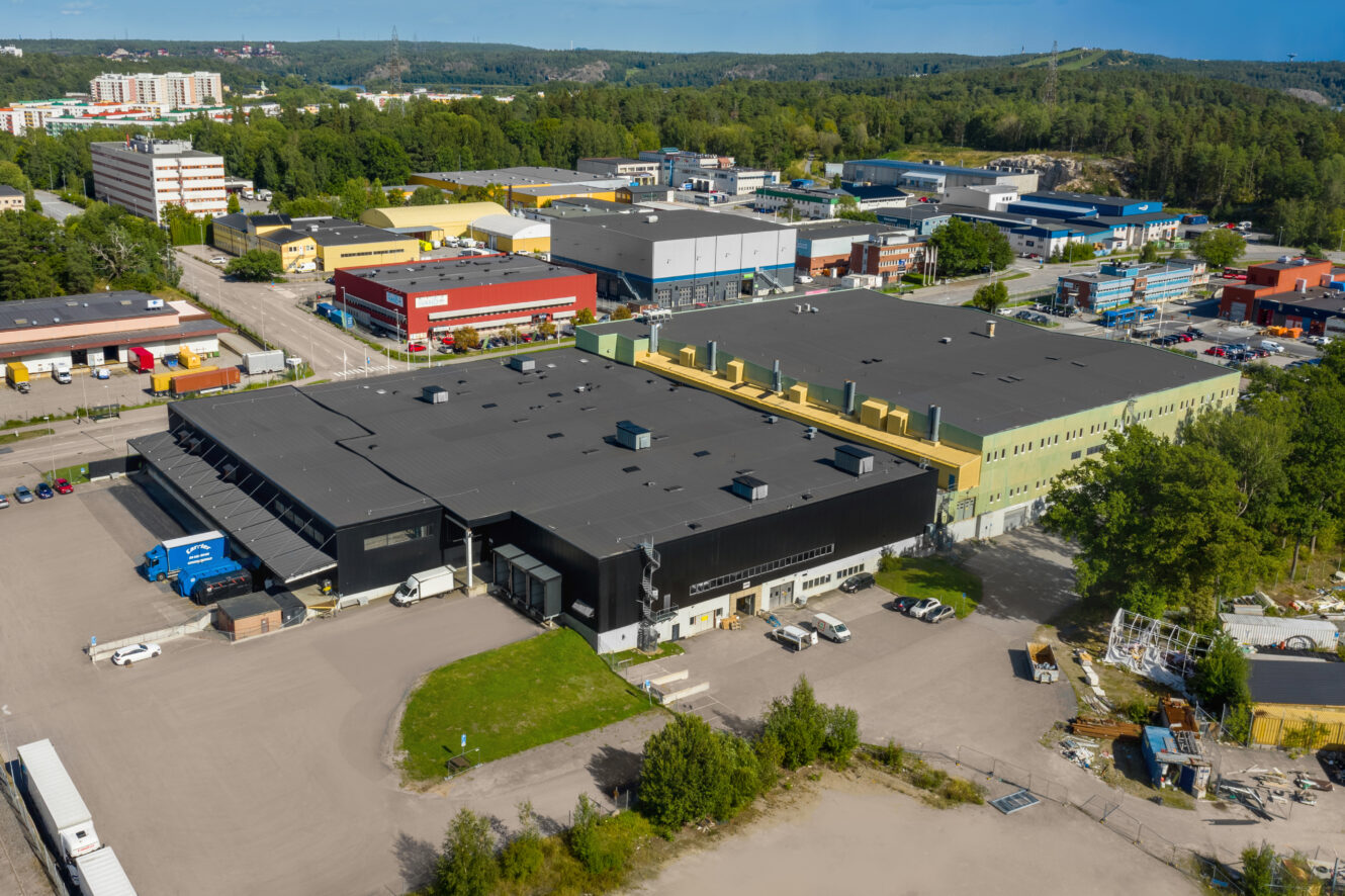 Flygfoto över ett industrikomplex med lagerlokaler, lastkajer, parkerade lastbilar och omgivande grönska i ett stadsområde.