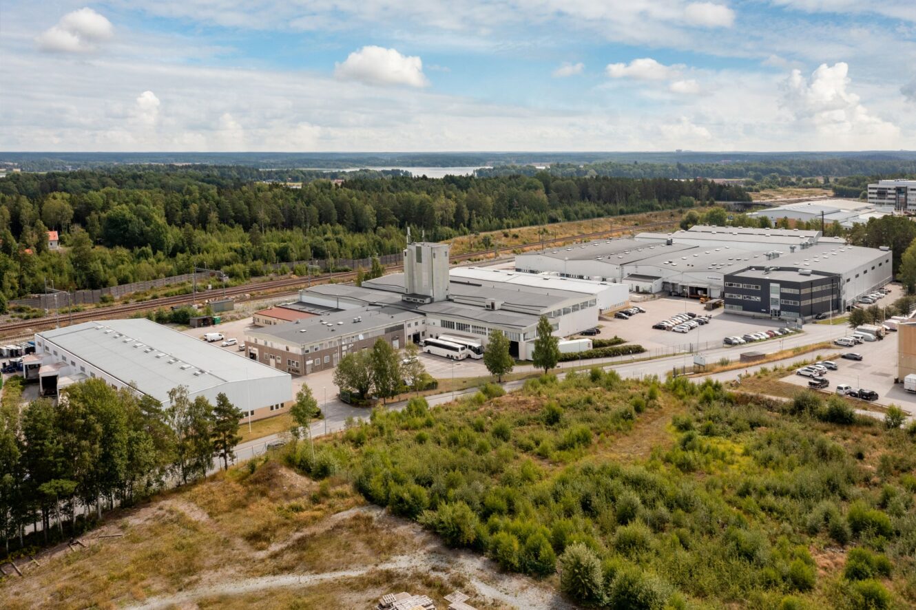 Flygfoto över ett stort industrikomplex med flera byggnader, parkeringsplatser och omgivande grönska under en delvis molnig himmel.