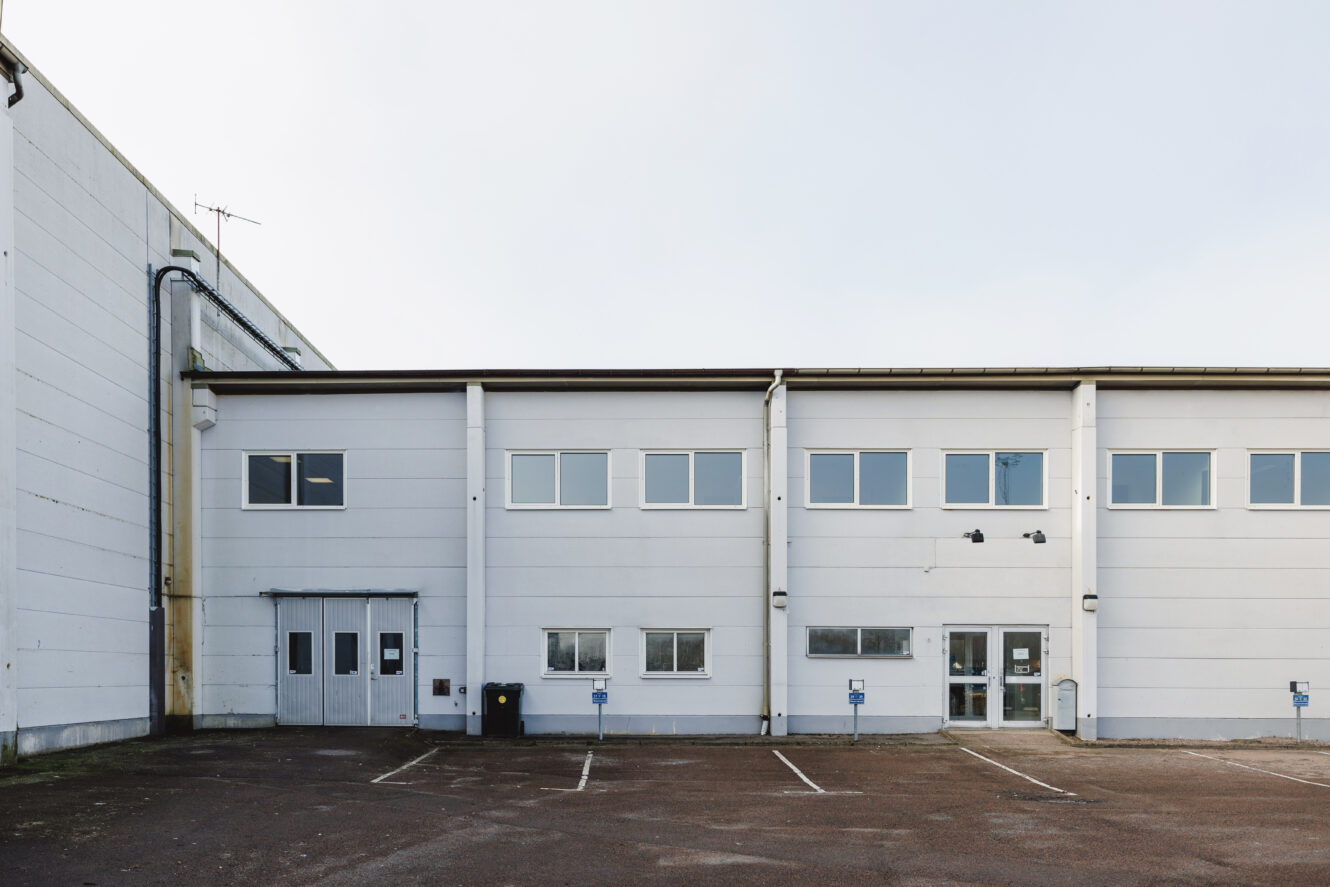 En vanlig, vit industribyggnad med flera fönster, dörrar och tomma parkeringsplatser framför sig en molnig dag.