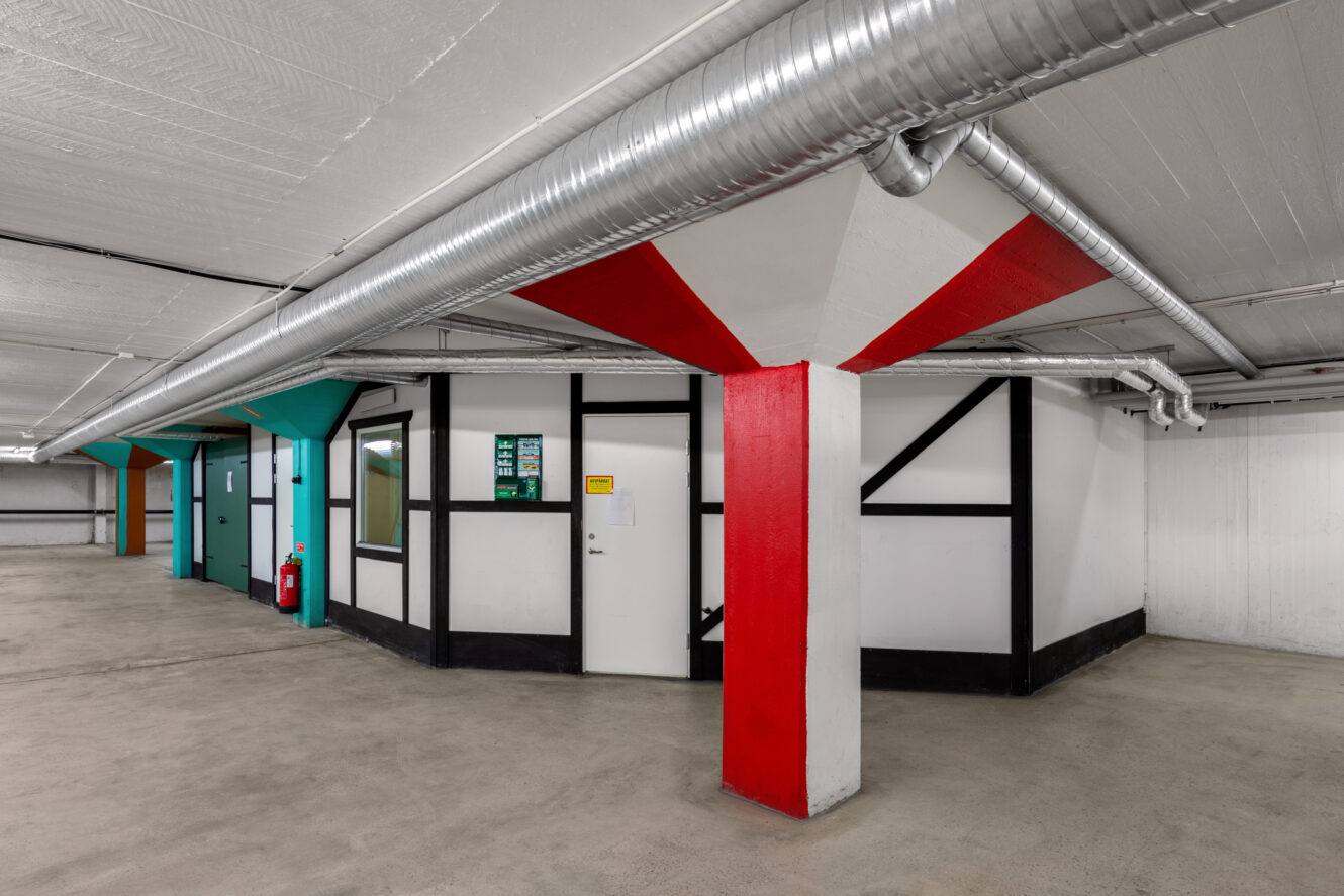 Ett rent, tomt parkeringsgarage med synliga rörledningar, vita väggar, geometriska färgaccenter på pelare och ett litet rum med dörrar och fönster.
