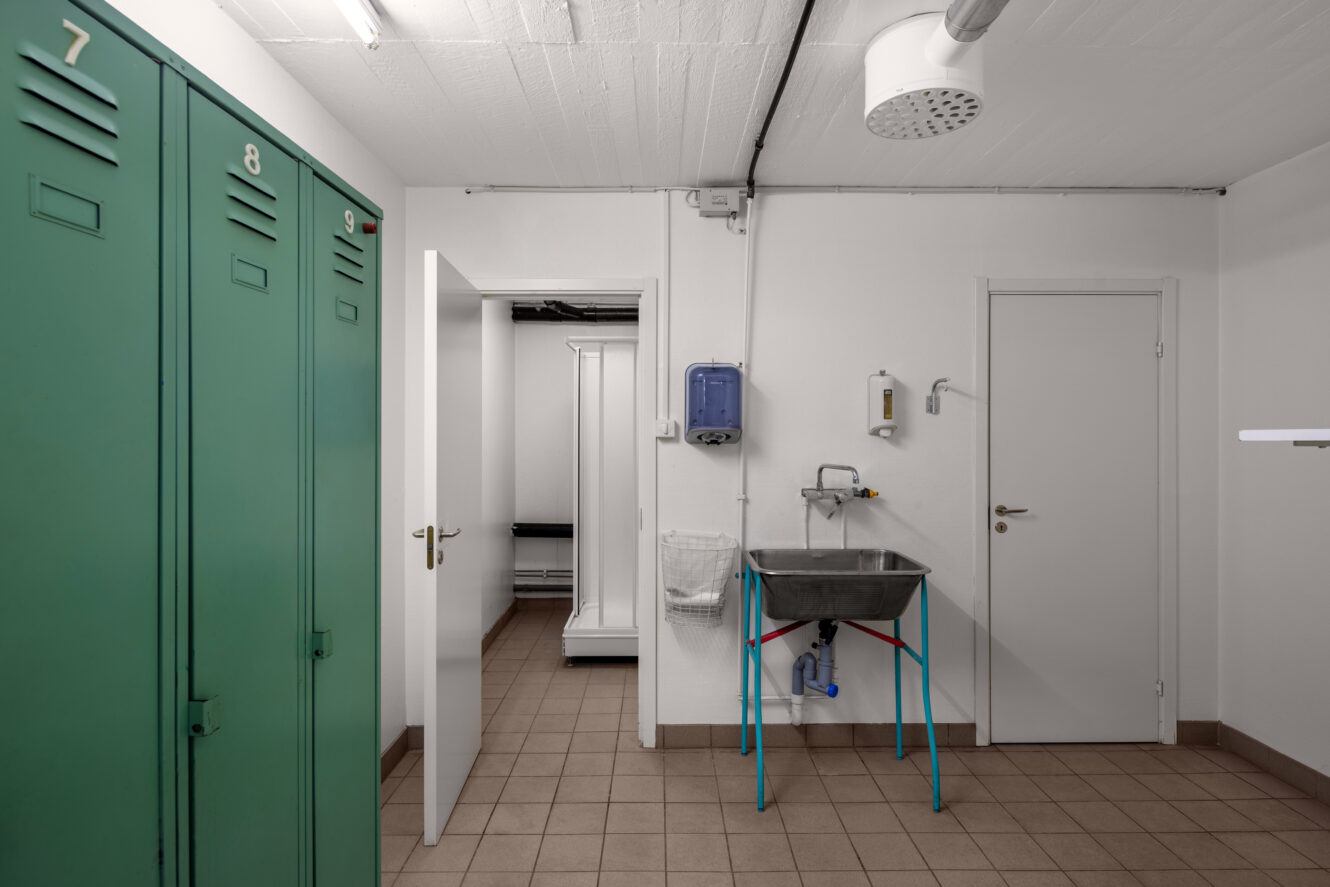 Industriellt omklädningsrum med gröna skåp, handfat i metall, tvålbehållare, klinkergolv och två vita dörrar som leder till angränsande rum.
