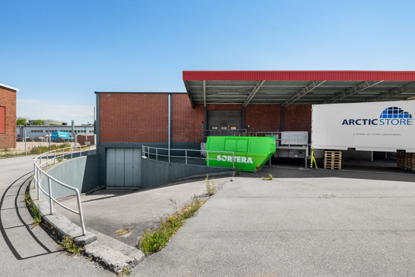 En grön container märkt SORTERA och en ArcticStore-container är placerade på en lastkaj utanför en industribyggnad i tegel under en klarblå himmel.