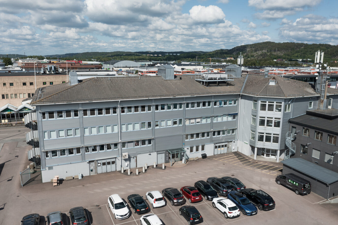 En stor grå industribyggnad med flera rader av fönster, en liten parkeringsplats framför och flera parkerade bilar. I bakgrunden syns industrianläggningar och kullar.