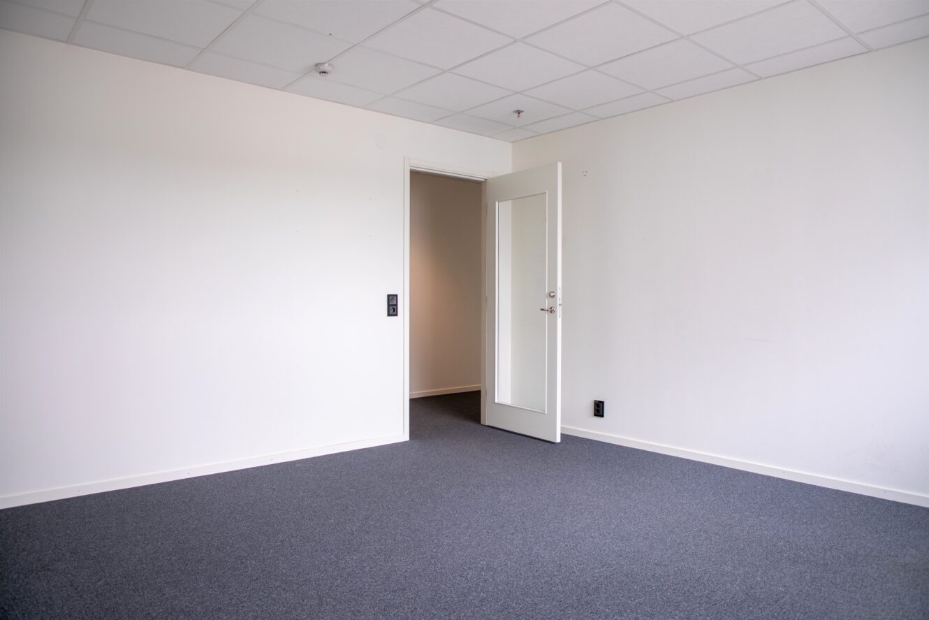 Tomt rum med vita väggar, en grå matta och ett vitt tak. En dörr med glaspanel är öppen och visar en korridor med liknande golv.