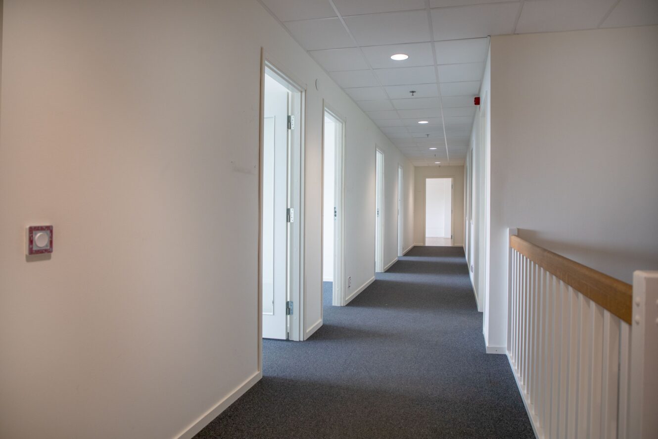 En ren, väl upplyst hall med vita väggar, grå heltäckningsmatta, flera öppna dörröppningar till vänster och ett vitt räcke till höger. Takplattor och infälld belysning är synliga.