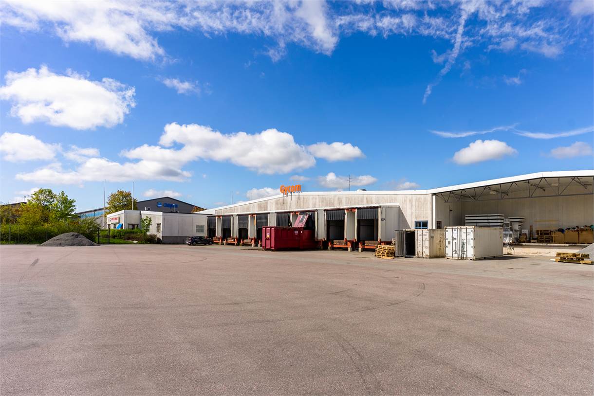 Ett stort industrilager med lastkajer, fraktcontainrar, en röd container och pallar under en blå himmel med spridda moln.