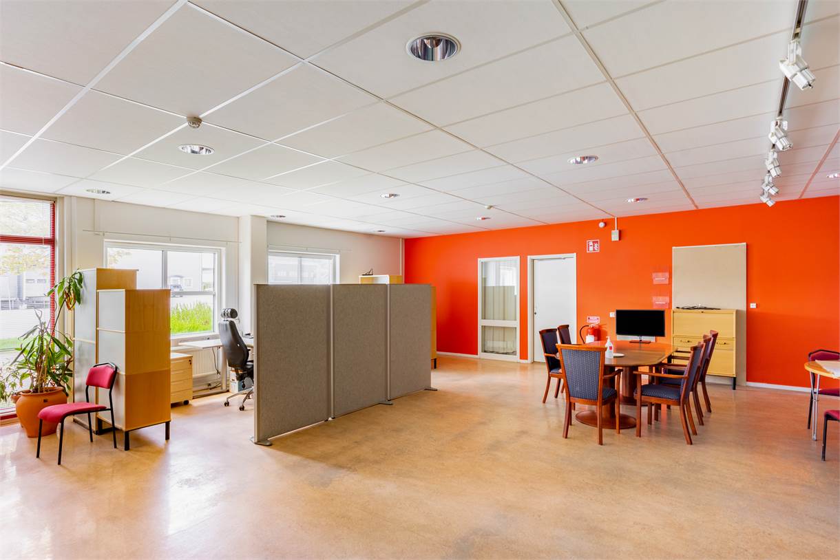 Modernt kontorsrum med orange accentvägg, avdelade skrivbord till vänster, ett runt mötesbord med stolar till höger och stora fönster som släpper in naturligt ljus.