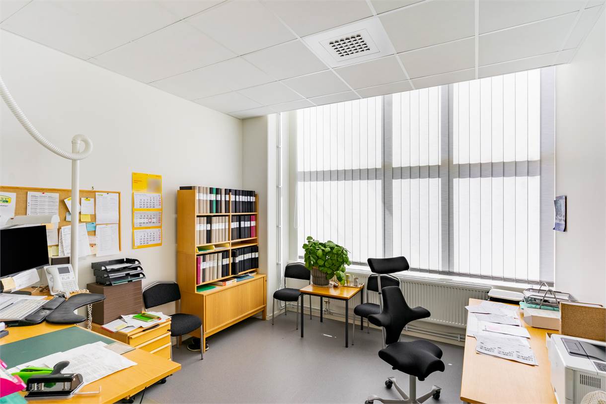 Ett väl upplyst kontor med skrivbord, stolar, en dator, hyllor fyllda med pärmar, krukväxt och papper utspridda på skrivborden. Stora fönster med vertikala persienner släpper in dagsljus.