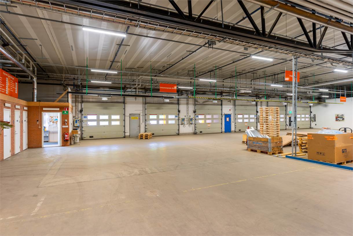 Rymlig lagerlokal med betonggolv, staplade pallar, stora garageportar och synliga takbjälkar under stark takbelysning.