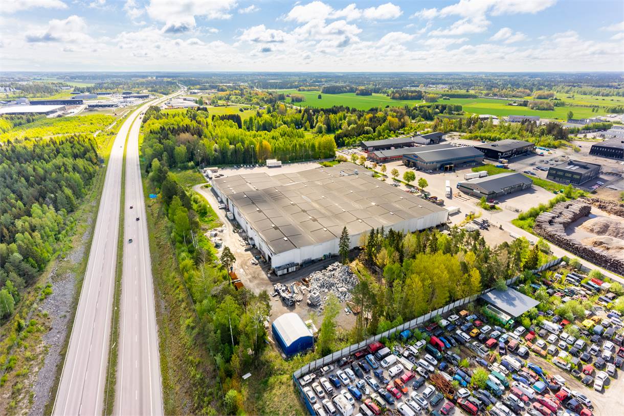 Flygfoto över en stor industribyggnad omgiven av träd, en parkeringsplats med bilar och en närliggande motorväg under en delvis molnig himmel.