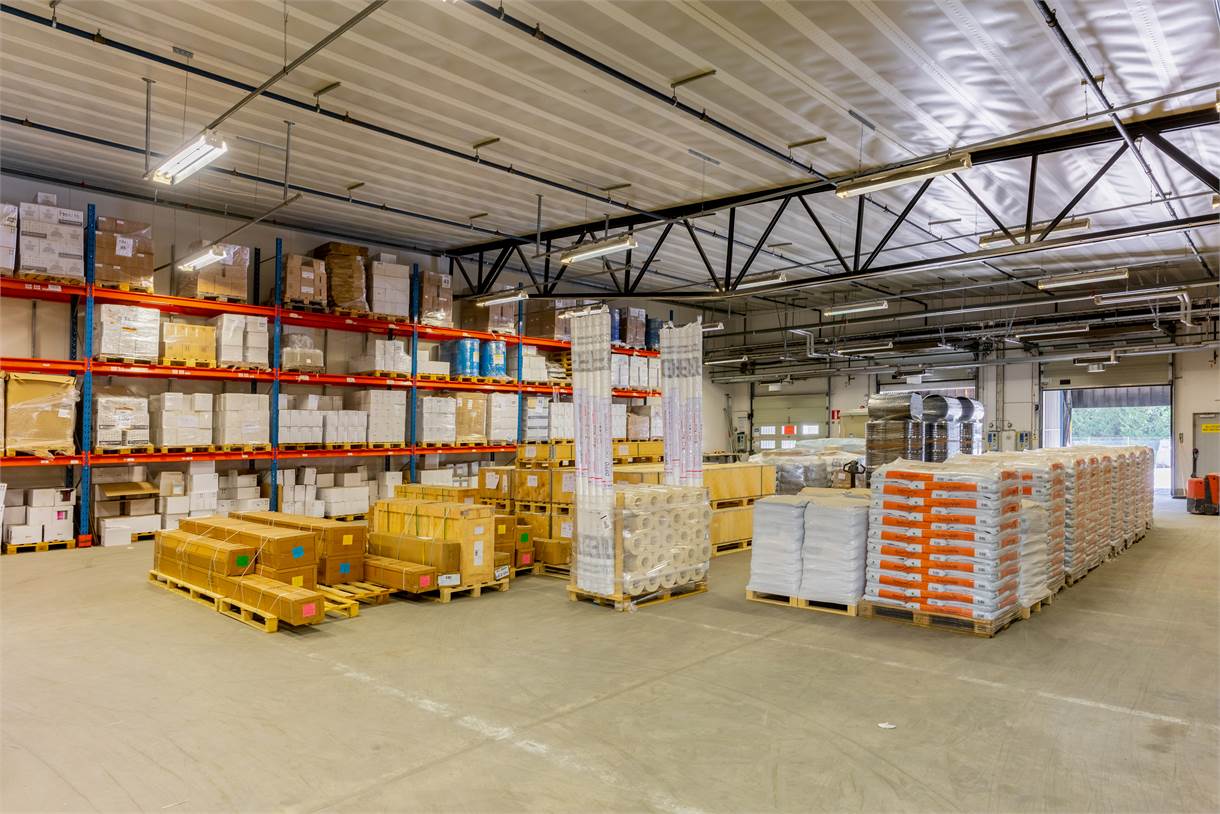 Rymlig lagerlokal med hyllor och pallar staplade med lådor och material, upplyst av taklampor.