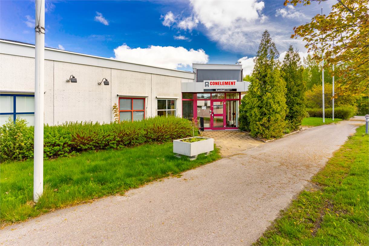 En vit envåningsbyggnad med en röd inramad entré, märkt GONELEMENT, omgiven av grönska och en asfalterad gångväg.