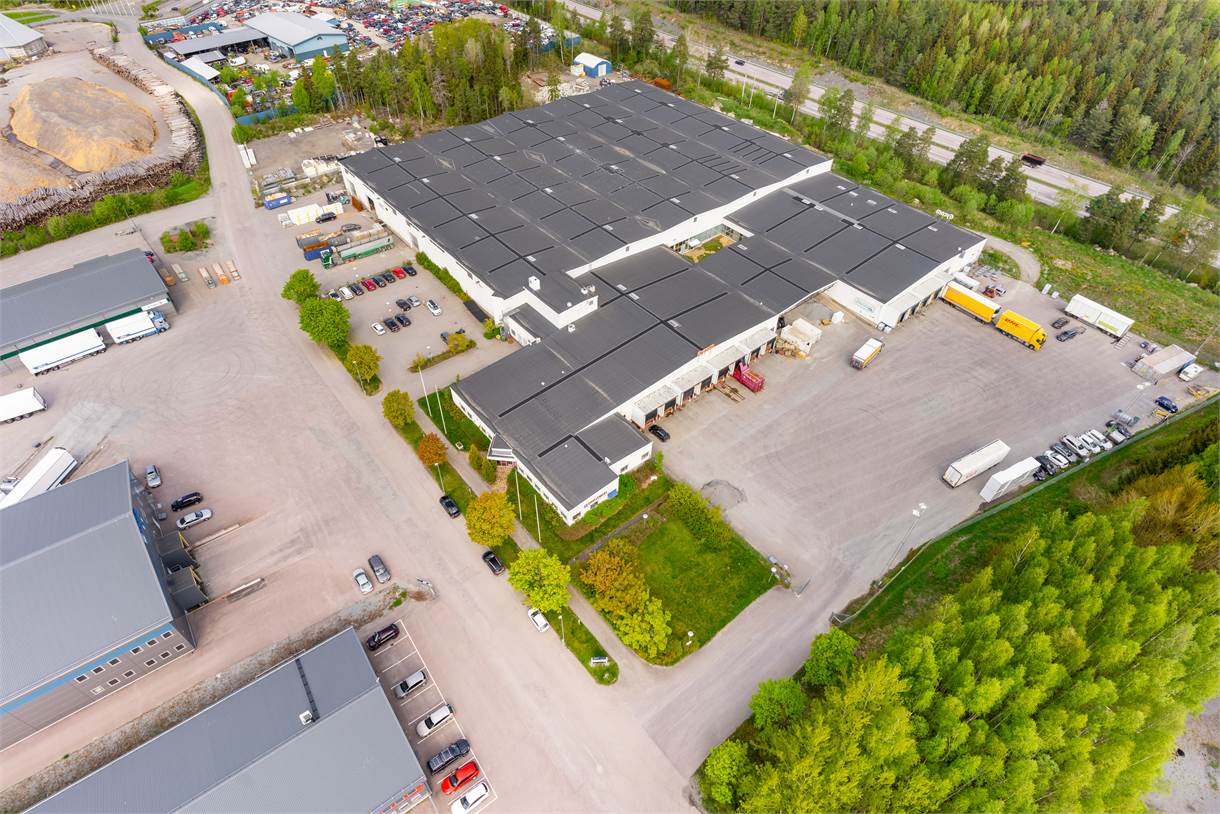 Flygfoto över ett stort industrilager med flera lastkajer, omgivet av lastbilar, bilar och grönska.