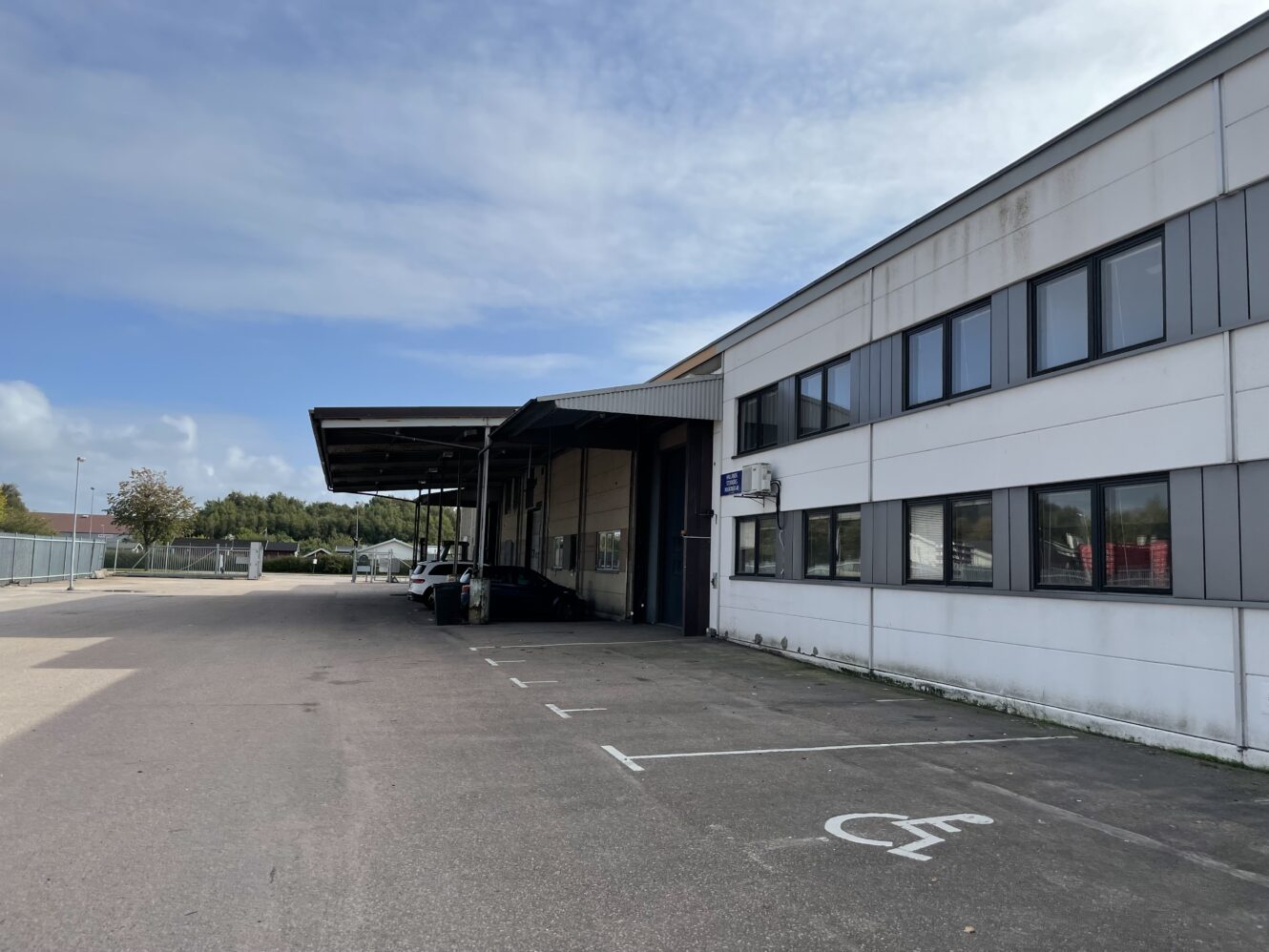 En utvändig vy av en kommersiell eller industriell byggnad med en täckt lastzon, parkerade bilar och tomma parkeringsplatser en solig dag.