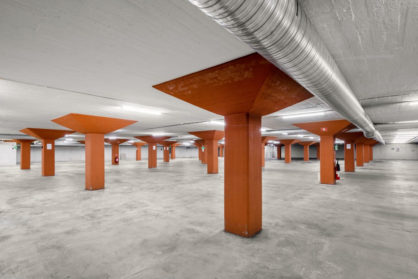 Stort, tomt underjordiskt parkeringsgarage med betonggolv och orangefärgade fyrkantiga pelare, upplyst av takbelysning och exponerade silverfärgade ventilationskanaler.